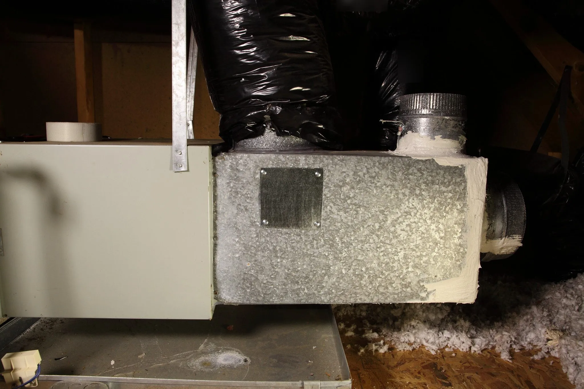 Air handler cabinet in an attic sitting in a galvanized drain pan, with a sheet metal plenum and black insulated flexible ducts
