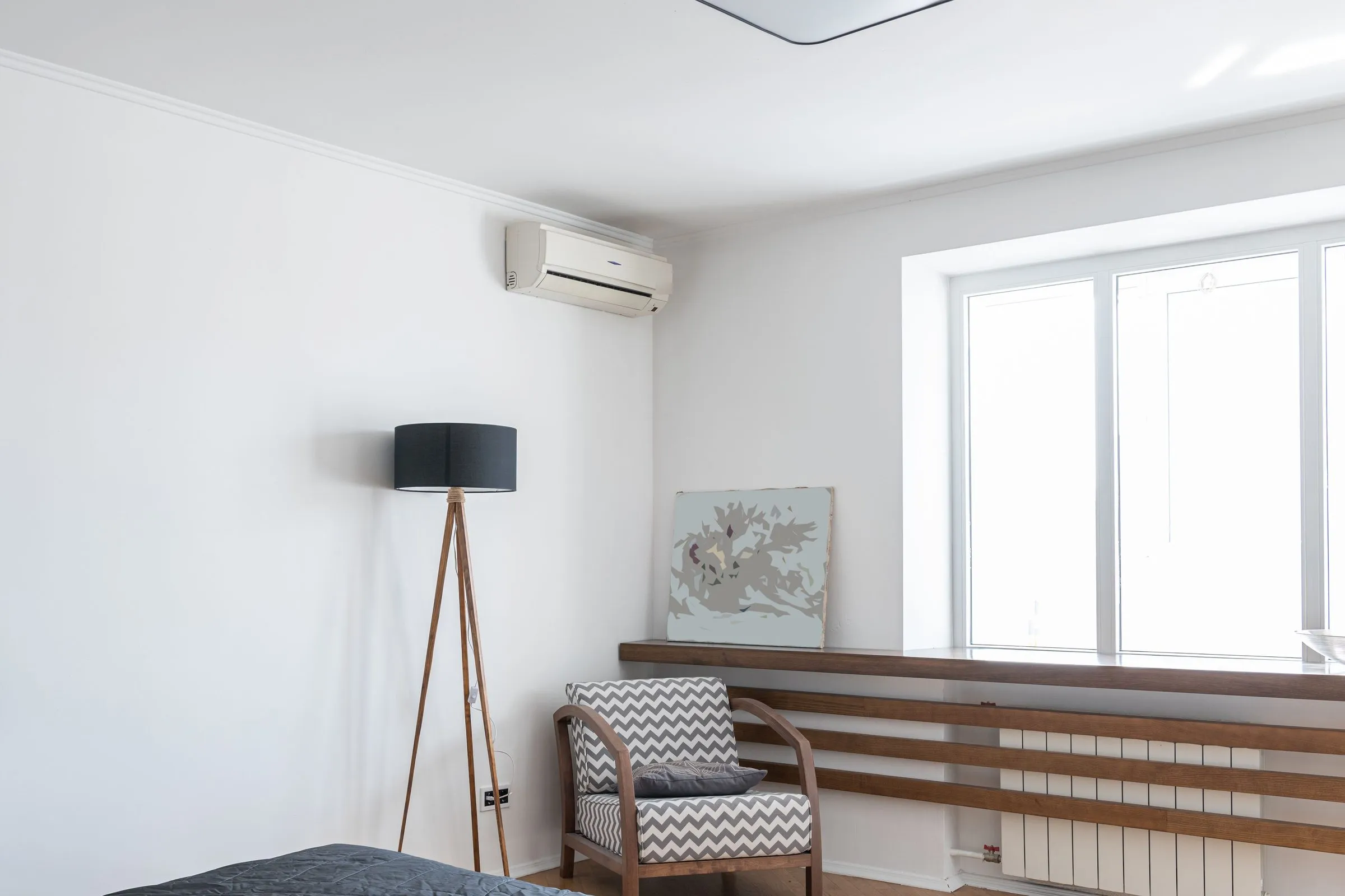 Bright bedroom with a ductless mini split indoor unit mounted high on the wall near the ceiling, above a floor lamp and an armchair
