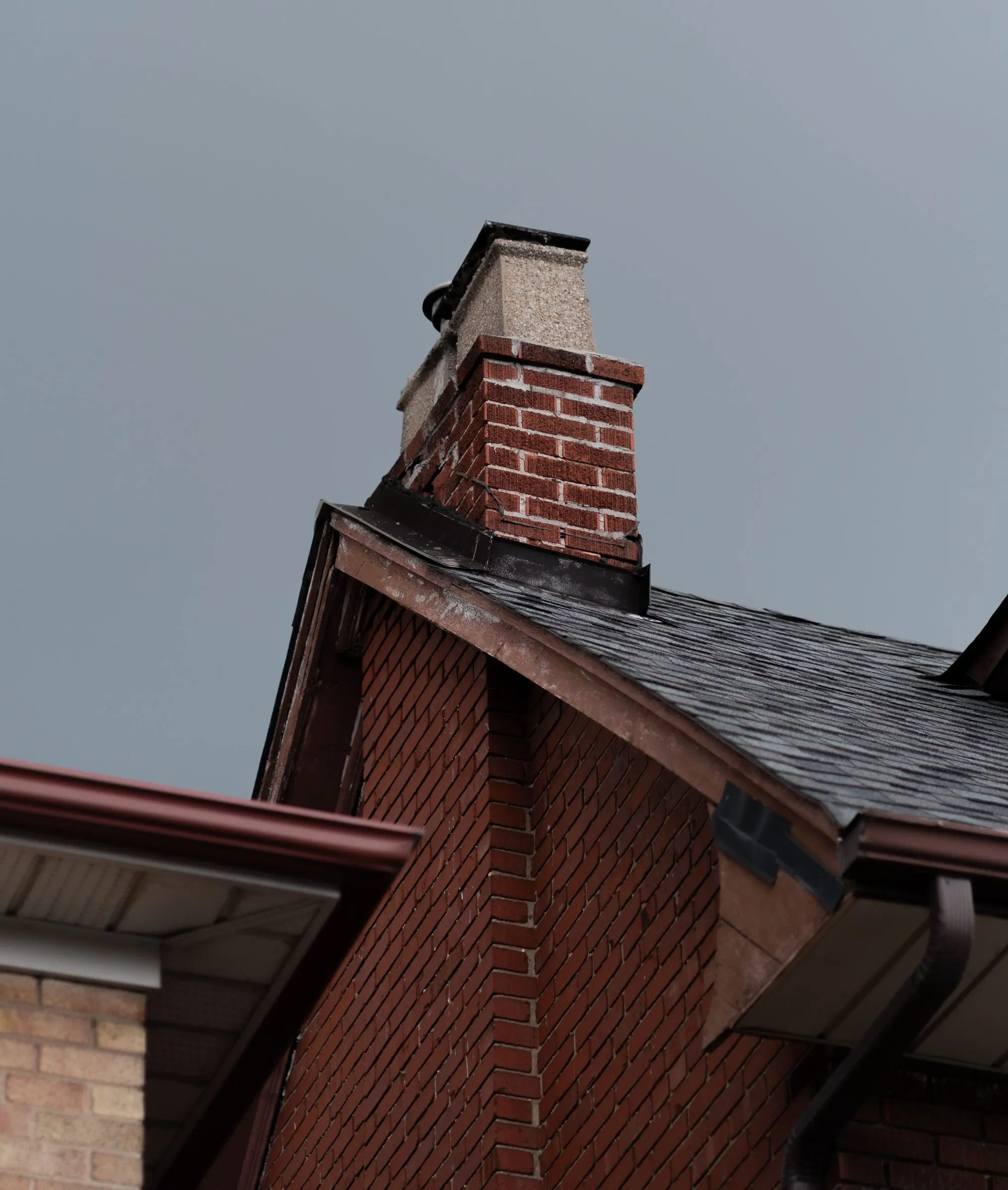 A brick chimney with a concrete cap rising from the shingled roof of a brick house under a gray sky
