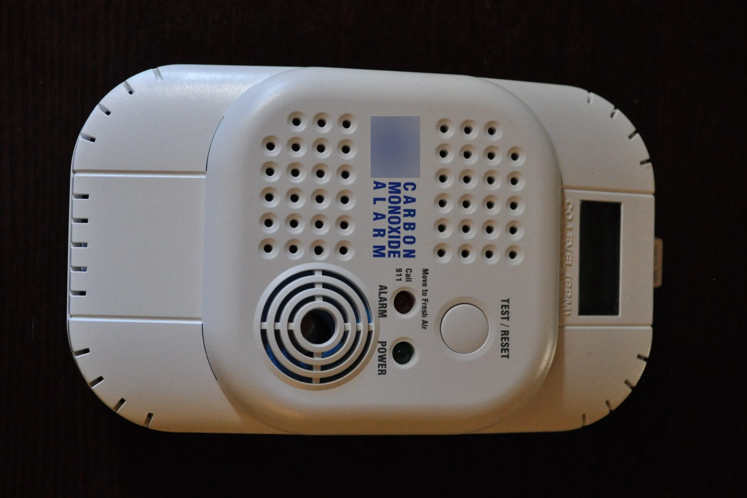 A white carbon monoxide alarm with a round test button, alarm and power lights and a display window, on a dark surface