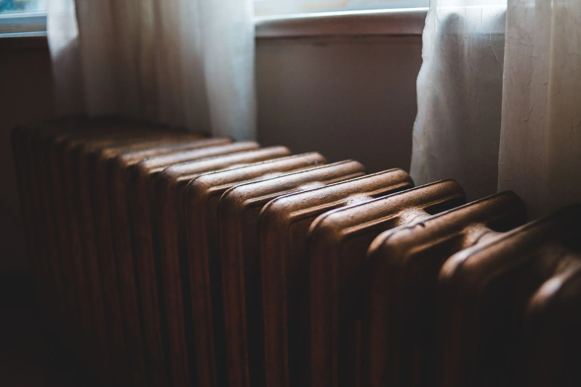 An old cast iron radiator under a window with sheer white curtains, in soft daylight
