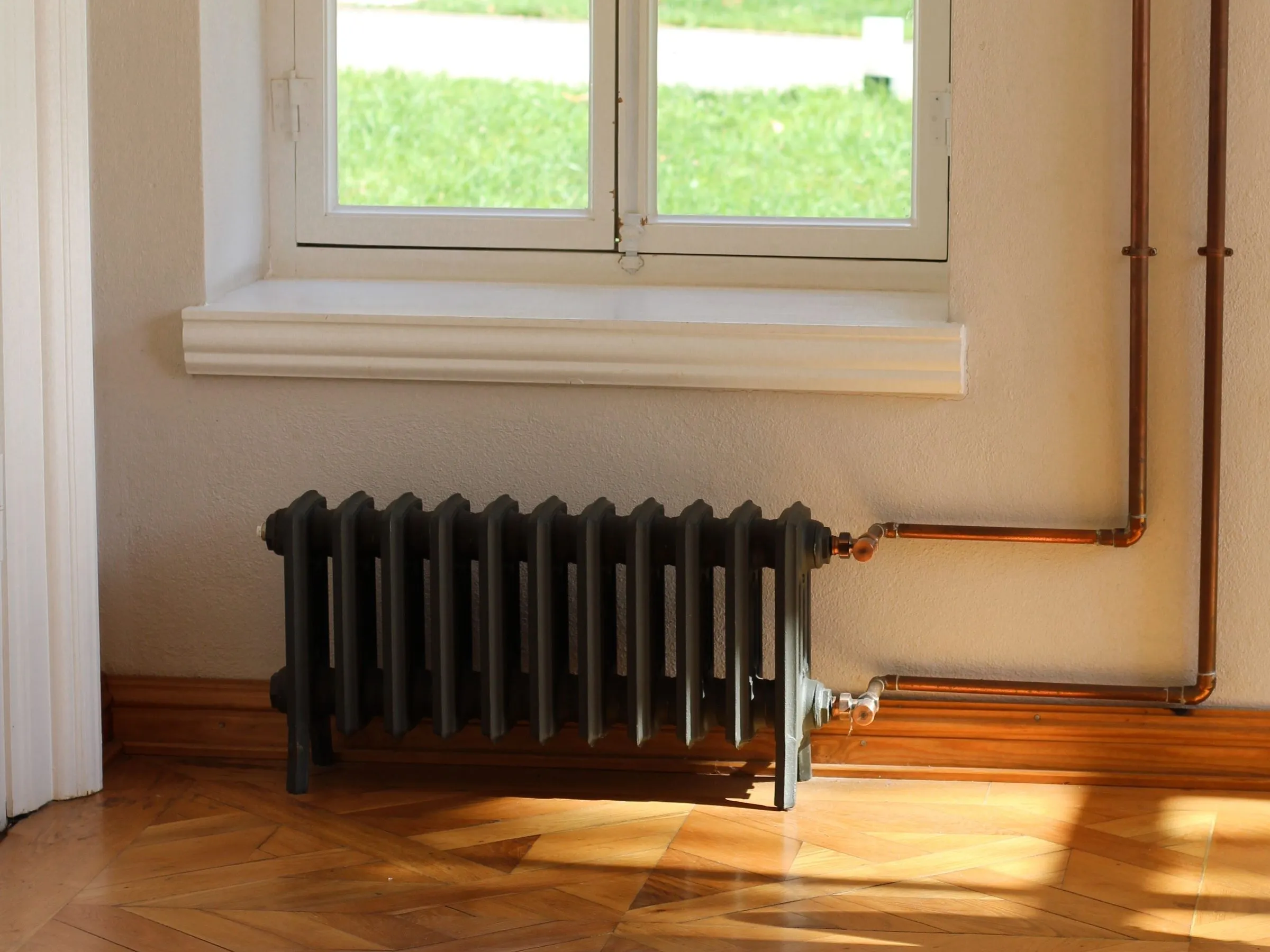 A dark cast iron column radiator under a sunny window, fed by two copper pipes with valves, on a parquet floor