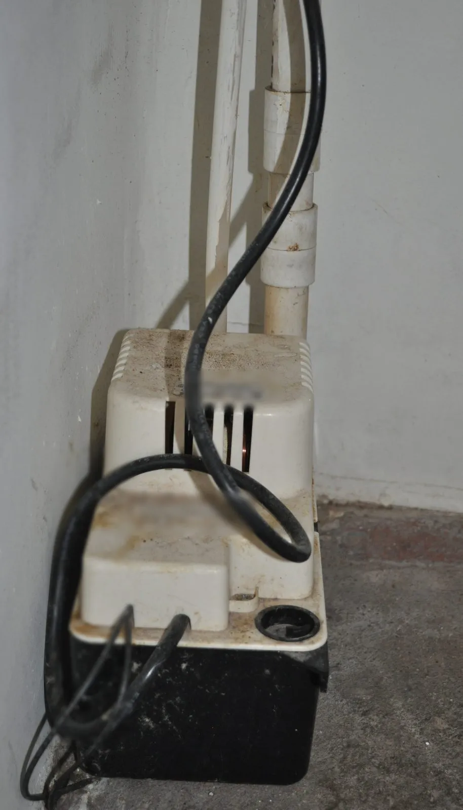 A small white condensate pump with a black reservoir on a basement floor, with white PVC drain pipes running down into it