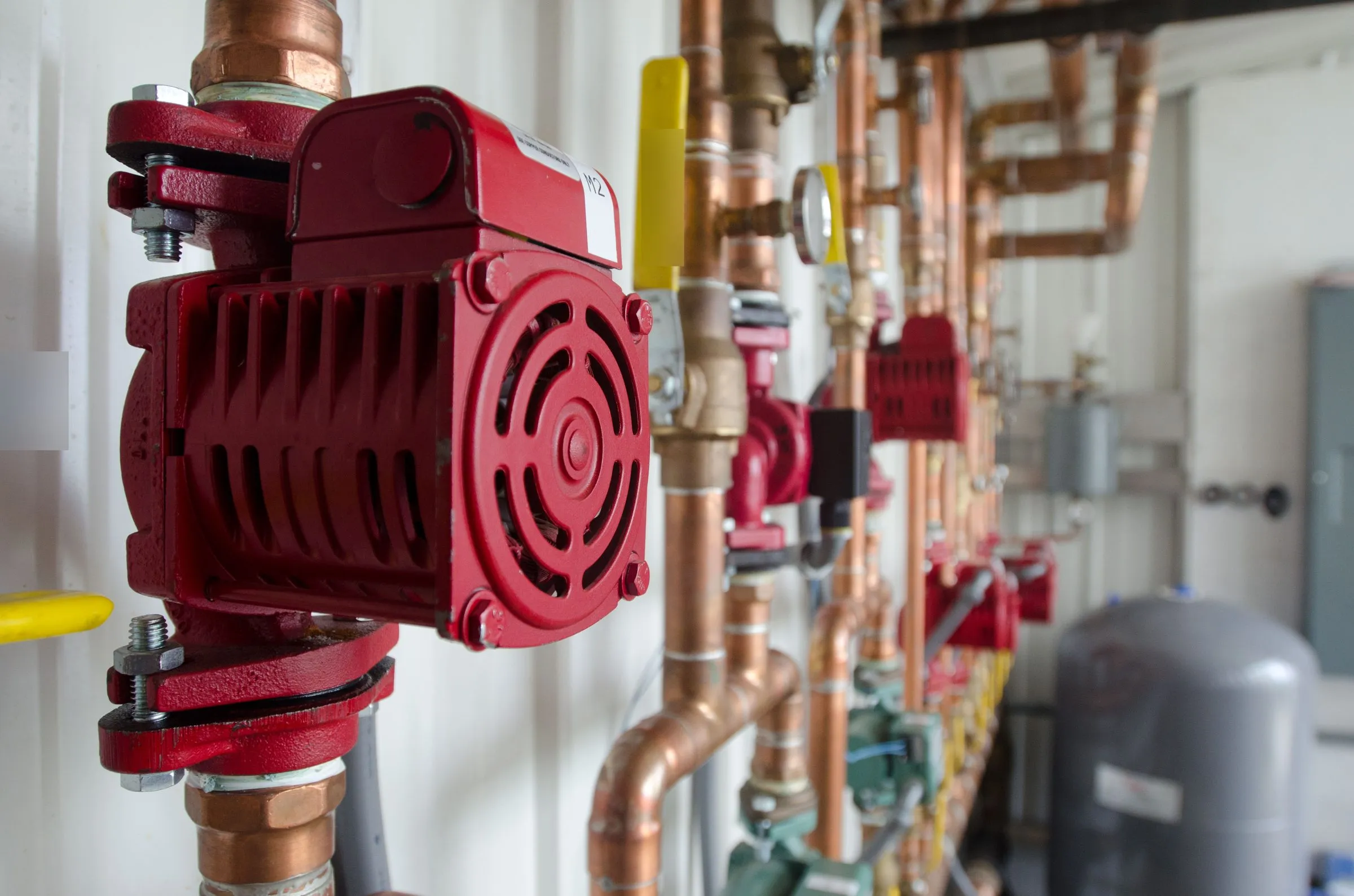 Copper hot water heating pipes in a boiler room, with red circulator pumps, valves and a gray expansion tank