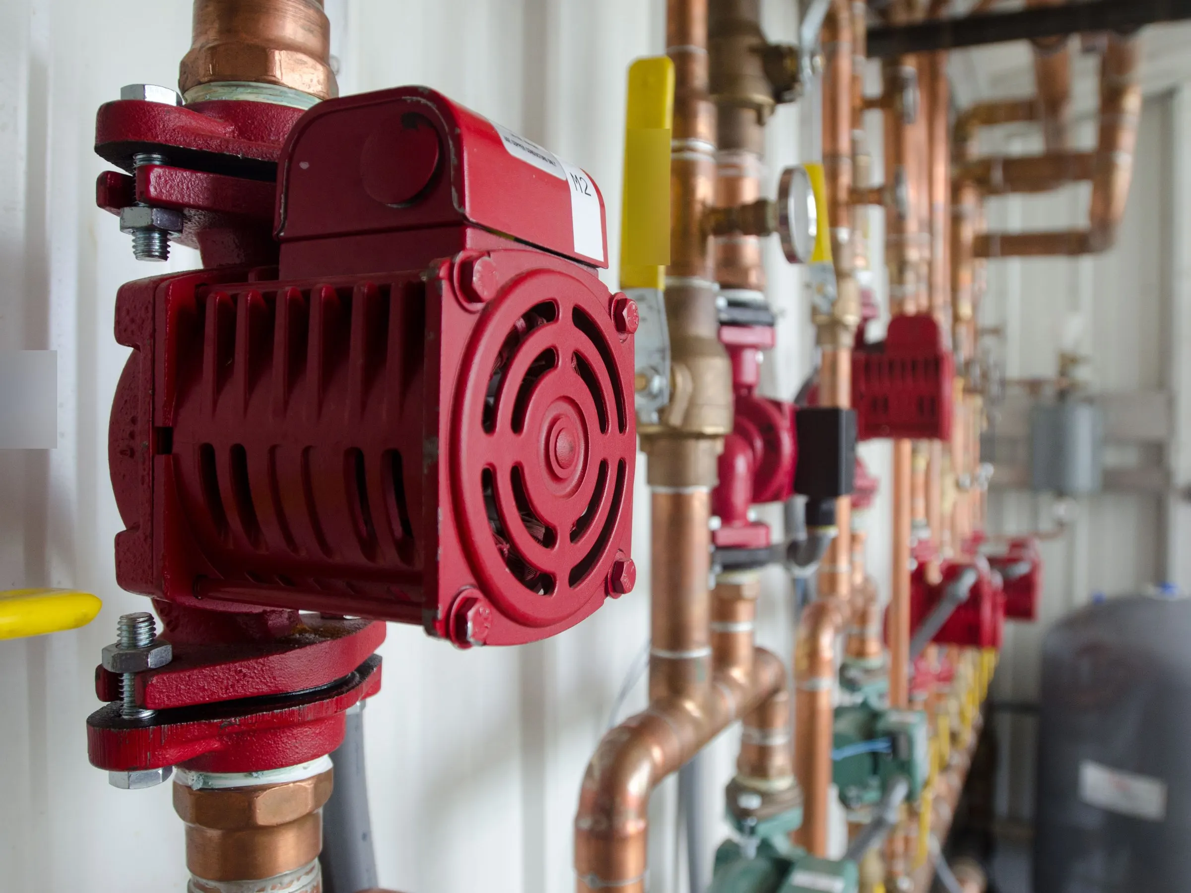 Copper hot water heating pipes in a boiler room, with red circulator pumps, valves and a gray expansion tank.