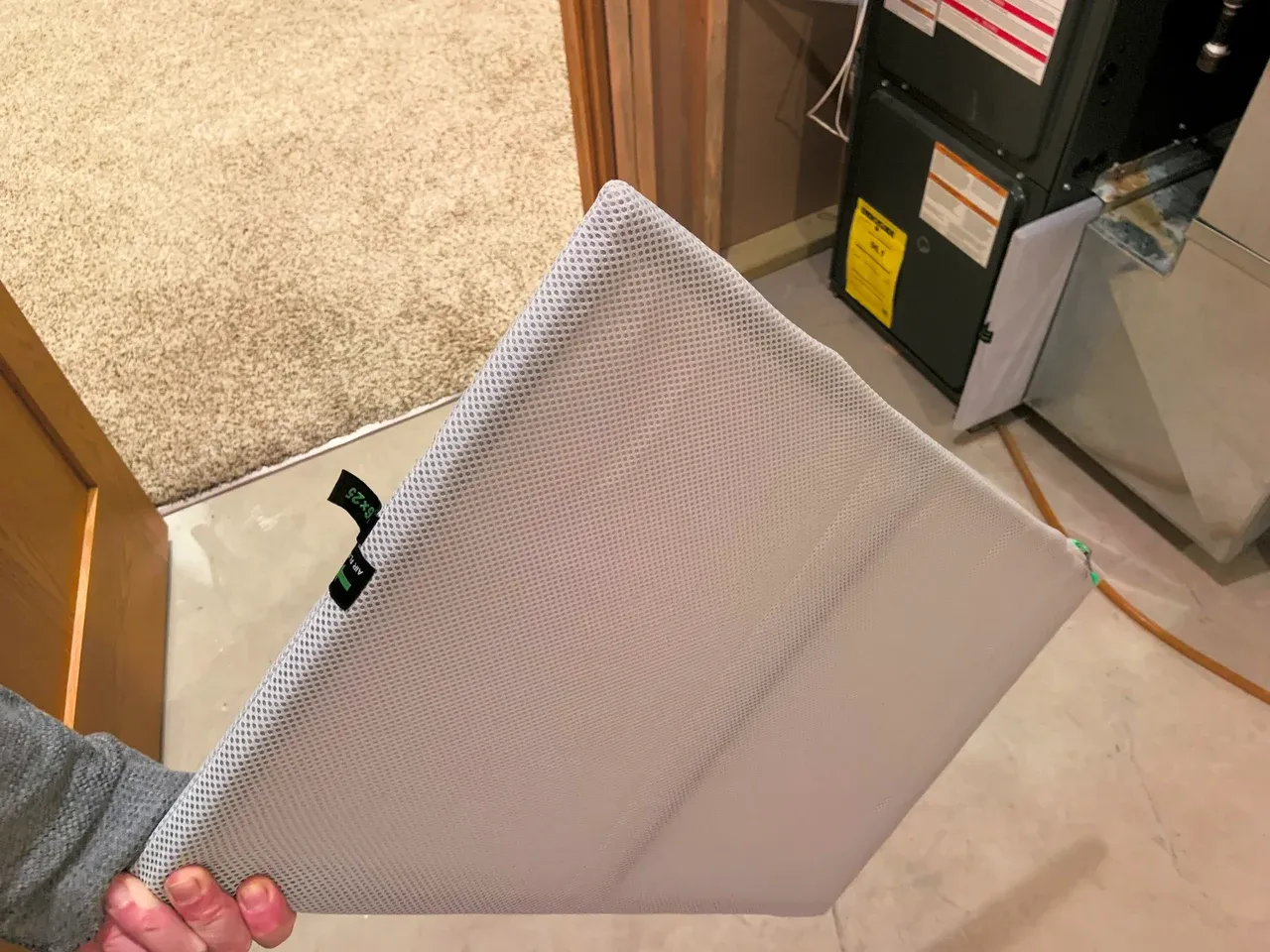 A person holding a rectangular air filter in a mesh cover next to a gas furnace in a basement