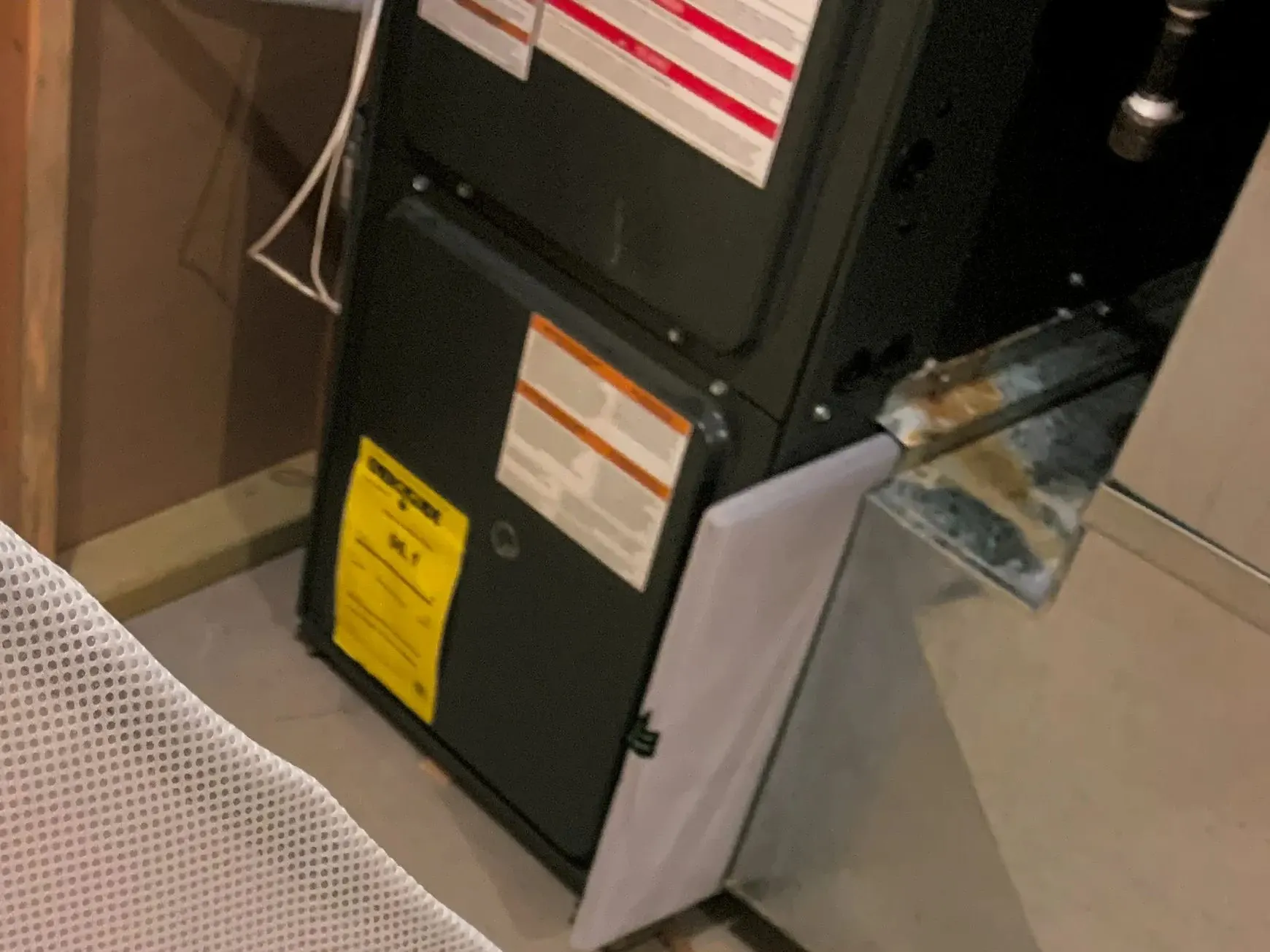 The lower cabinet of a gas furnace in a basement, with its yellow EnergyGuide label and warning labels on the door.
