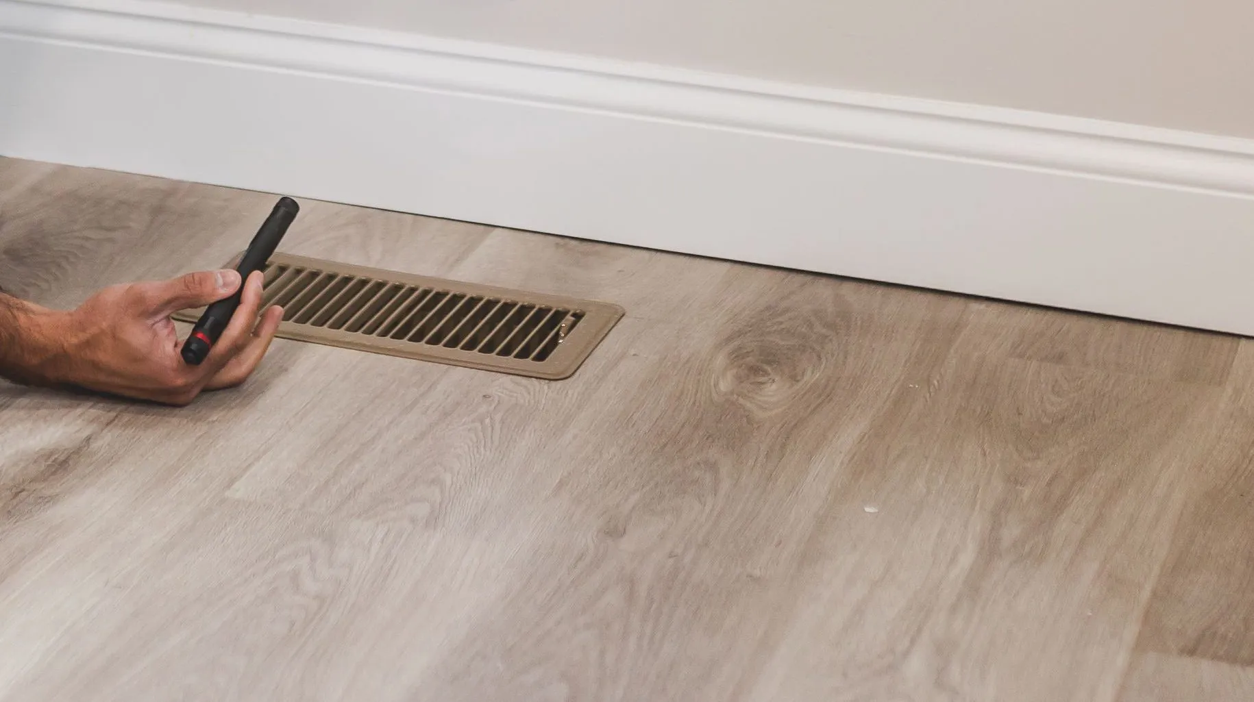 A hand holding a small penlight beside a metal floor heating register on a wood look floor next to a white baseboard