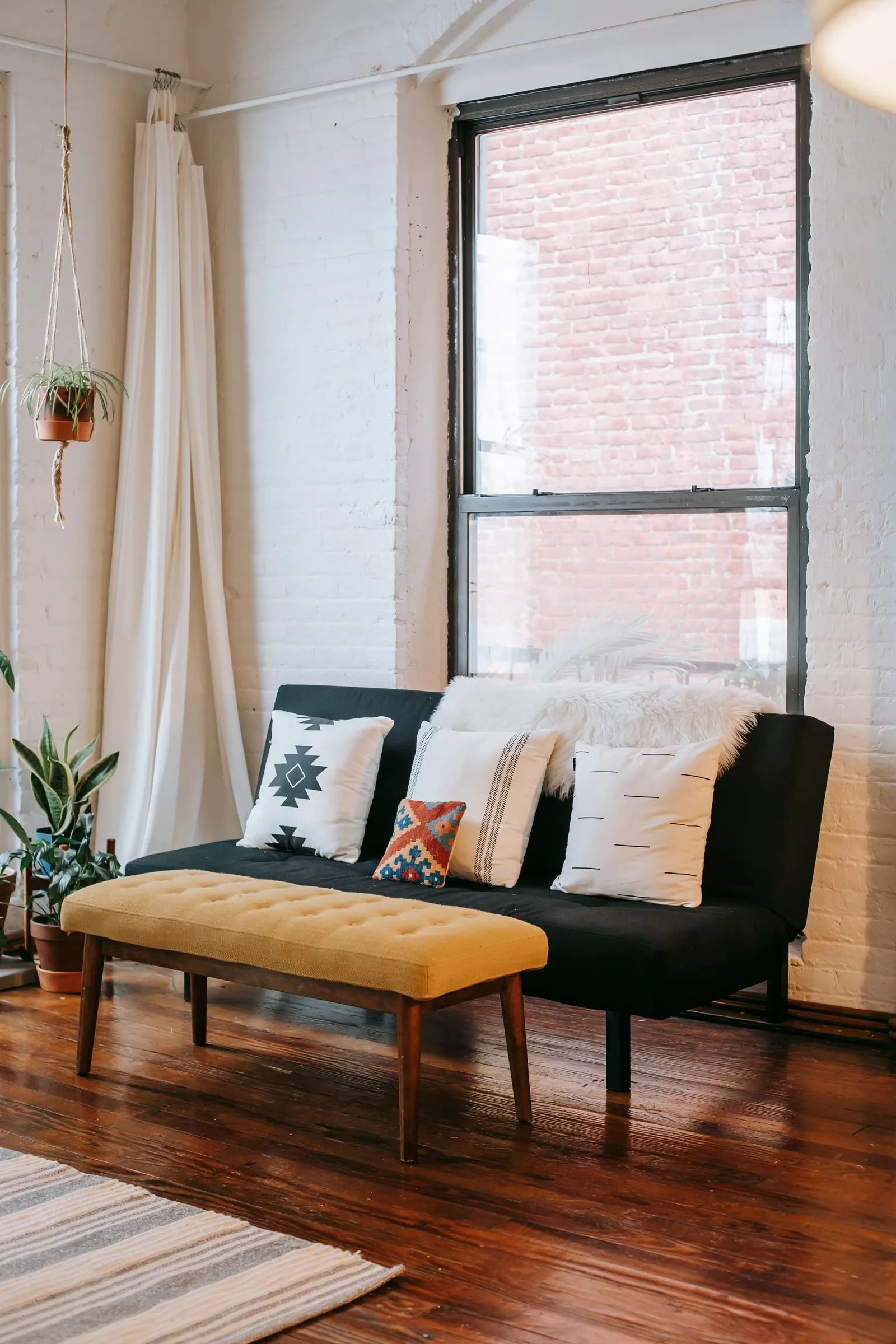 A bright living room with a black sofa and cushions under a tall window, a wooden bench in front and a hanging plant beside the curtain