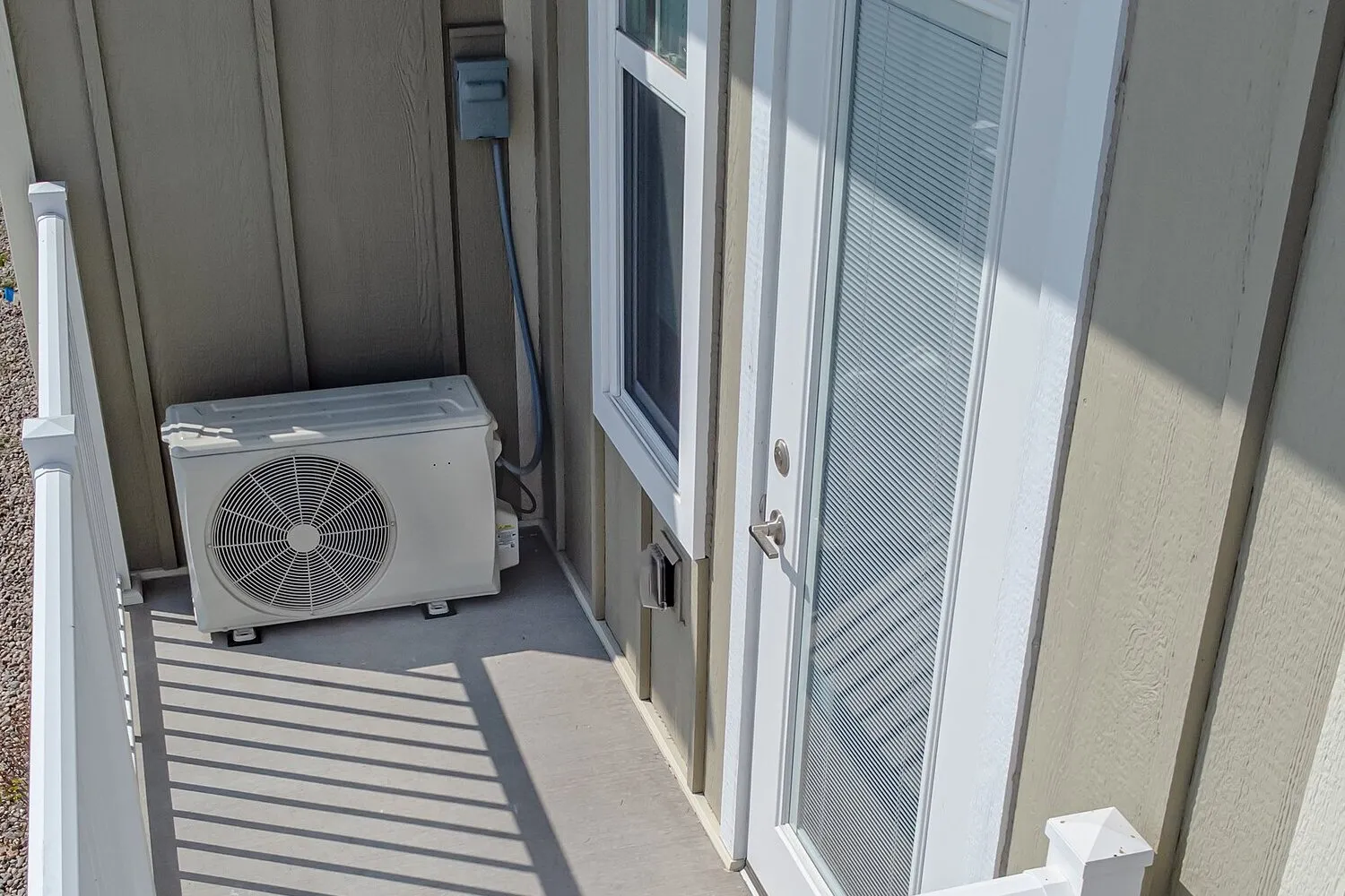 Outdoor heat pump unit on the floor of a small balcony beside a door, against the wall of a house