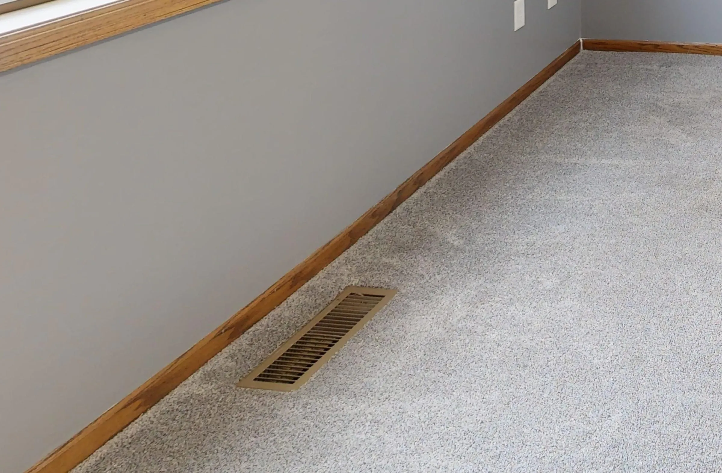 Metal floor register set into gray carpet beside a wood baseboard, where warm air enters the room in a forced air system