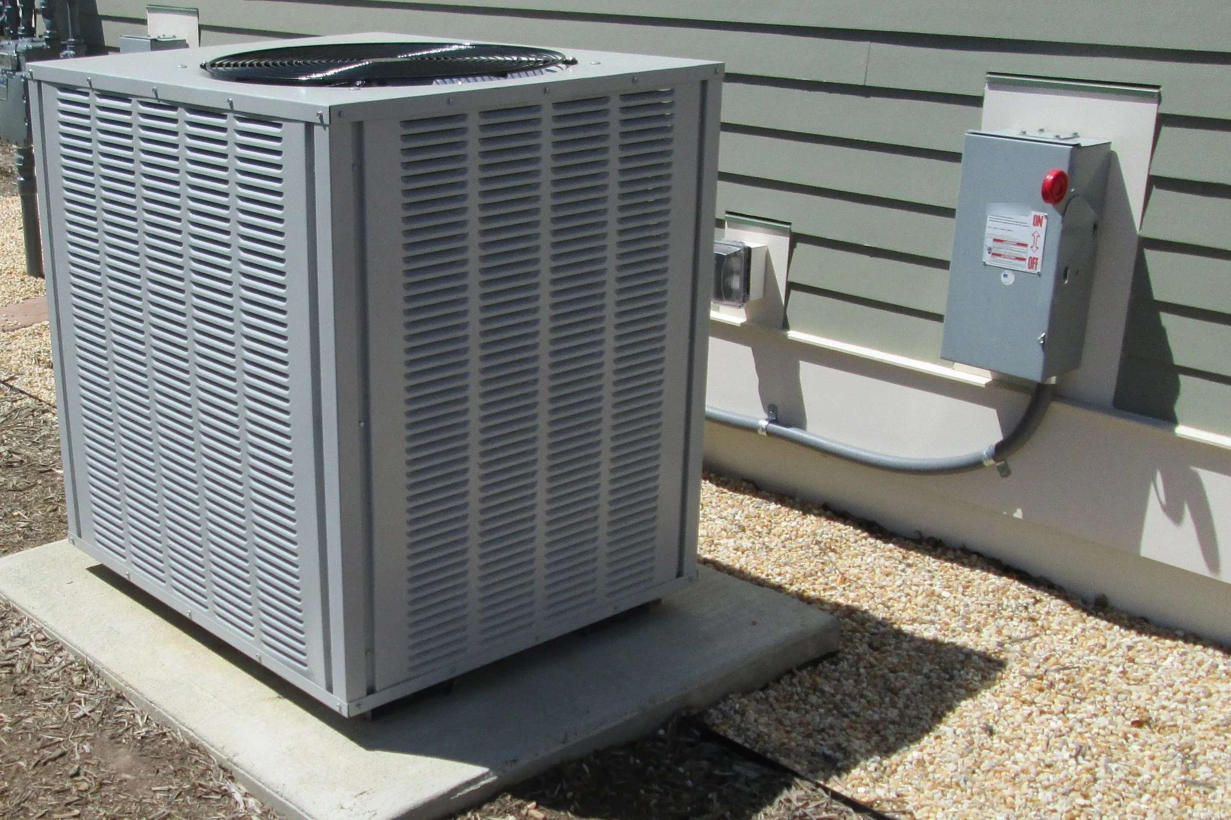 Outdoor central air conditioner on a concrete pad beside a house wall, with a fan grille on top, louvered side panels and a shutoff box