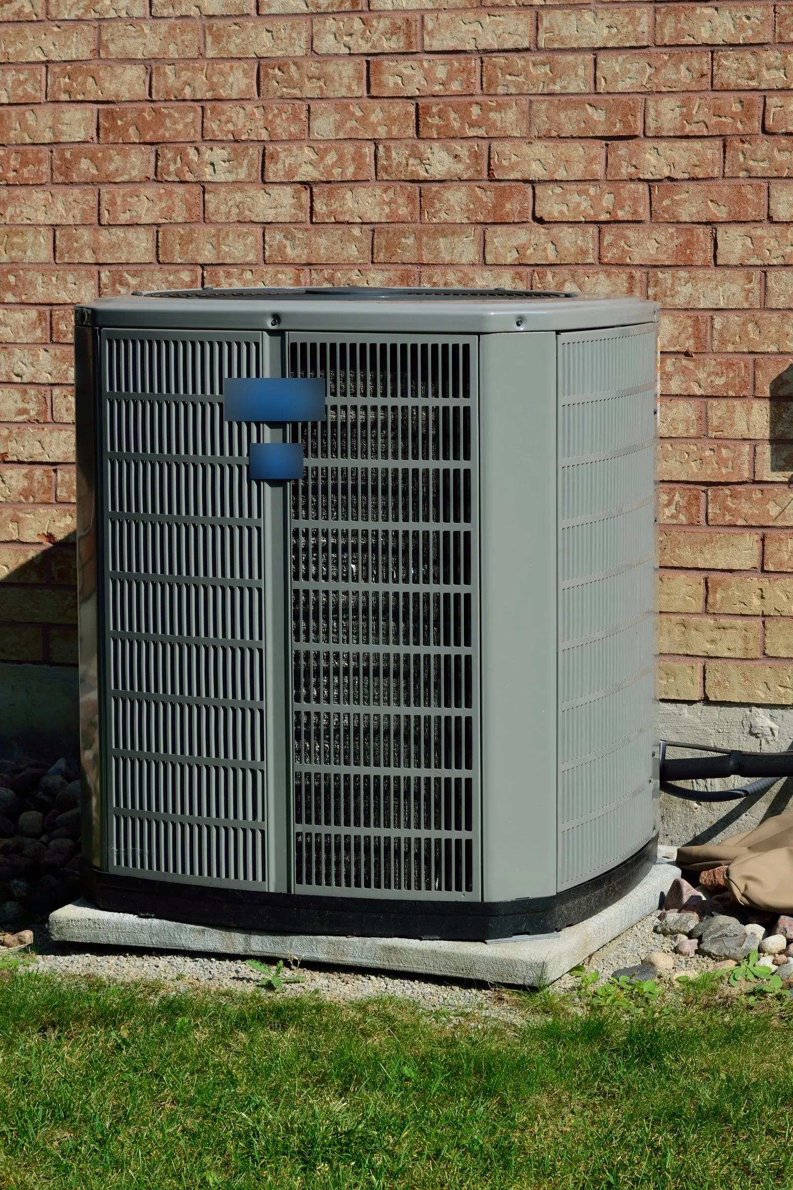 Outdoor air conditioner condenser with slotted gray panels on a concrete pad beside a brick house wall