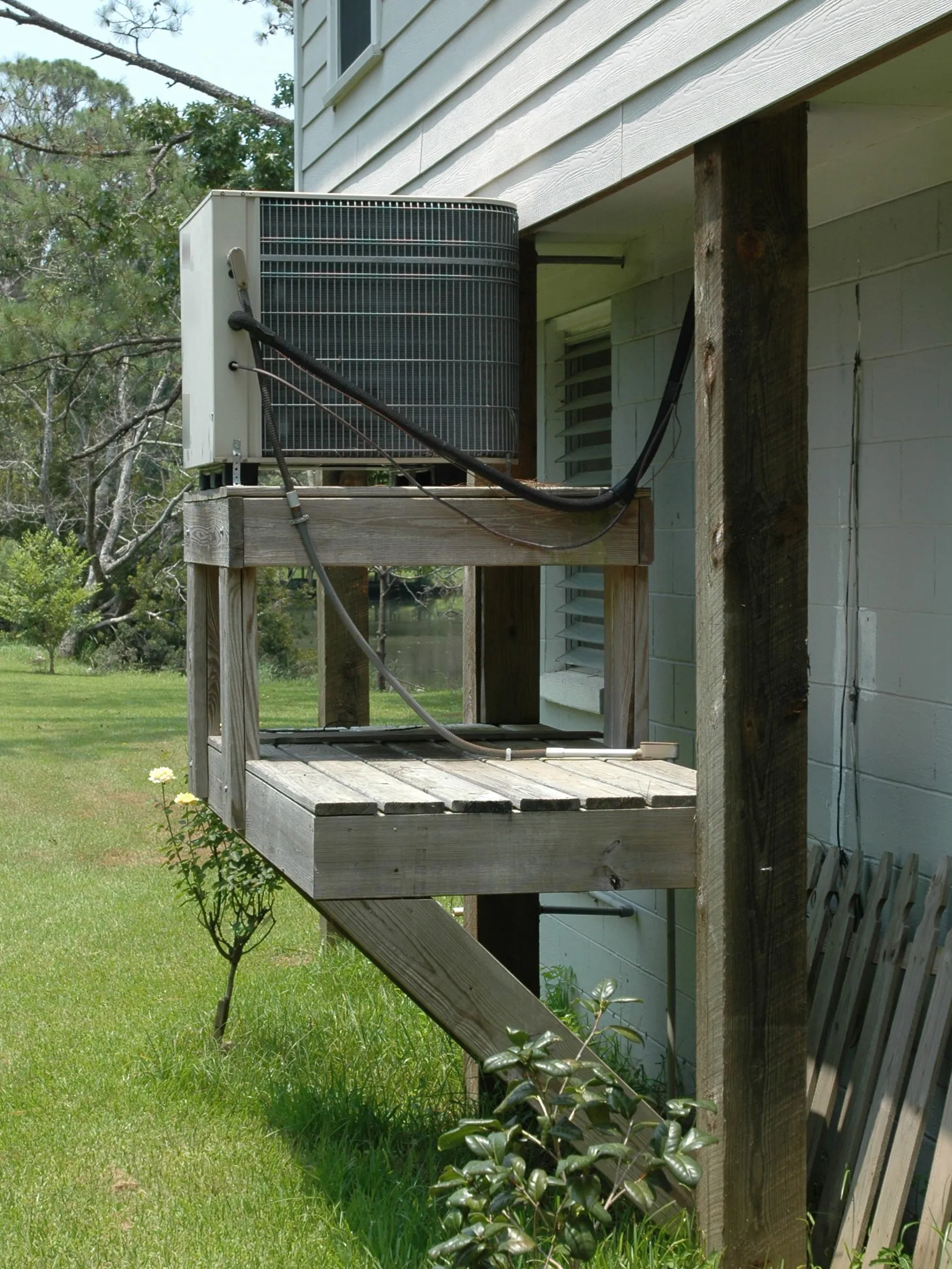 Outdoor air conditioner condenser raised on a wooden stand beside a house near the water, with refrigerant lines running to the wall