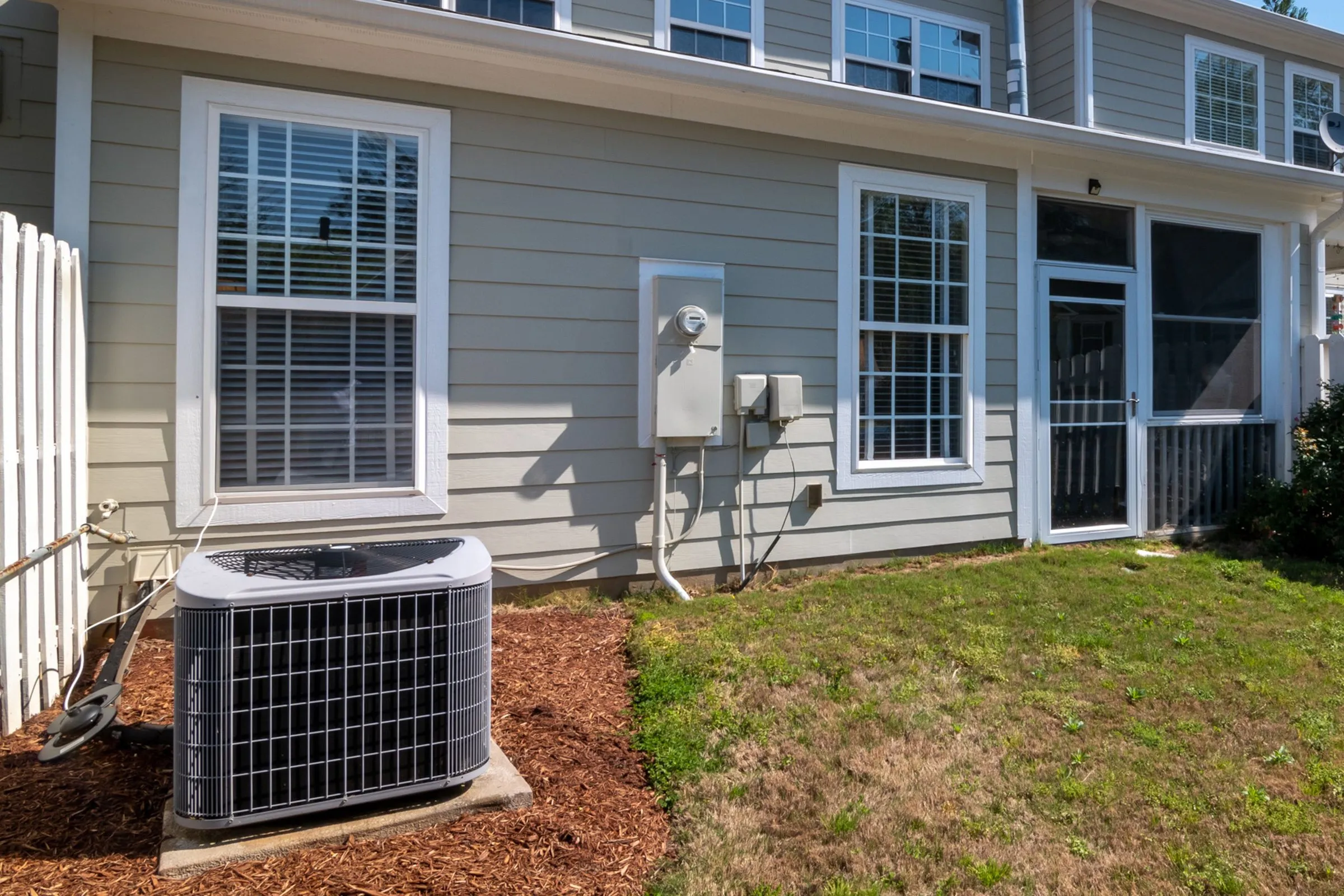 Outdoor unit of a split heating and cooling system on a concrete pad in a mulch bed behind a two-story house with a small lawn