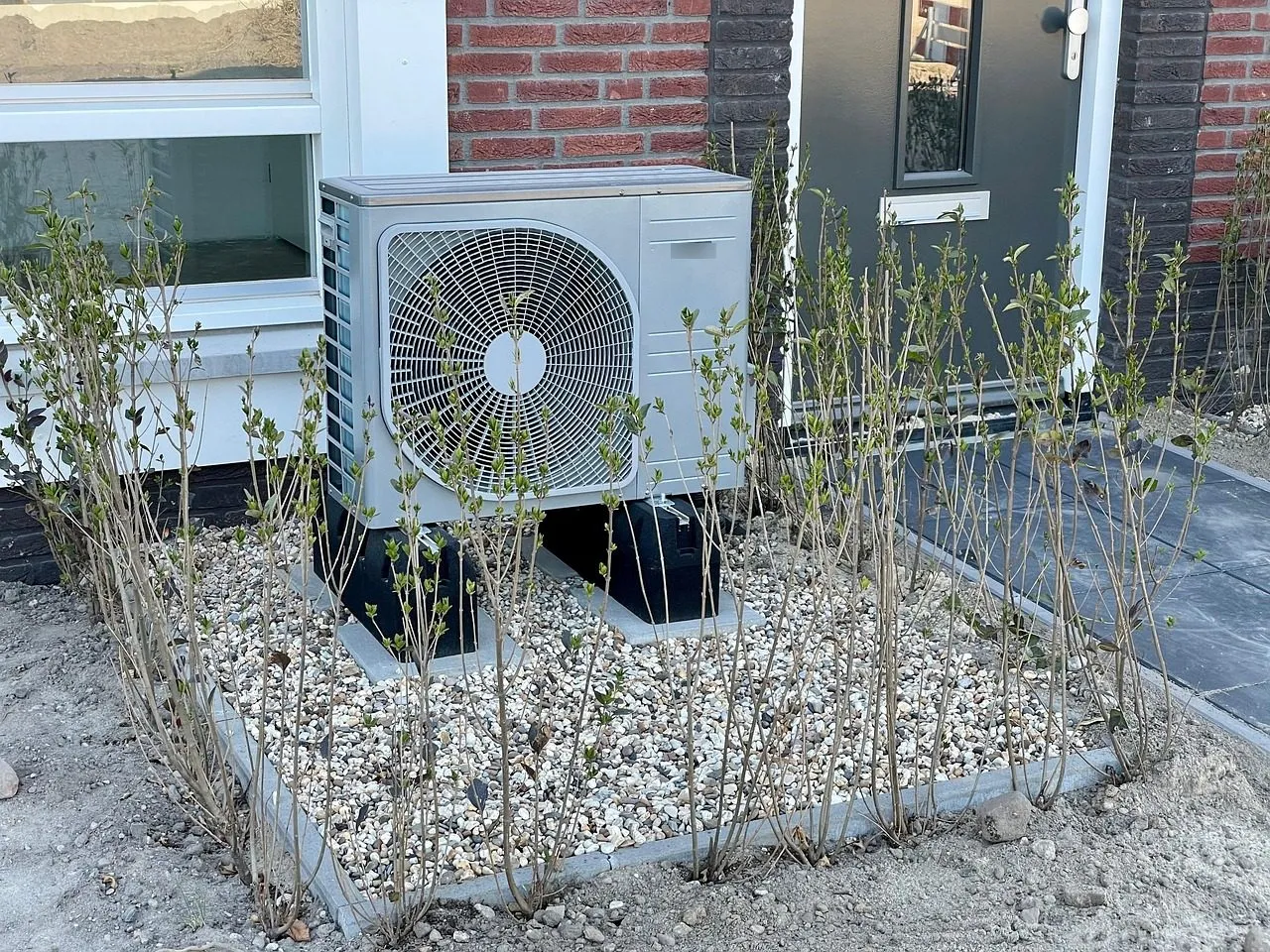 Outdoor heat pump unit raised off the ground on plastic feet over a gravel bed beside a brick house, with young shrubs around it