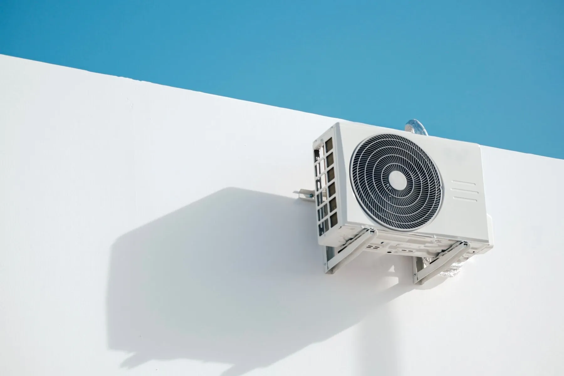 Outdoor unit of a ductless mini split mounted on a wall bracket high on an exterior wall against a blue sky