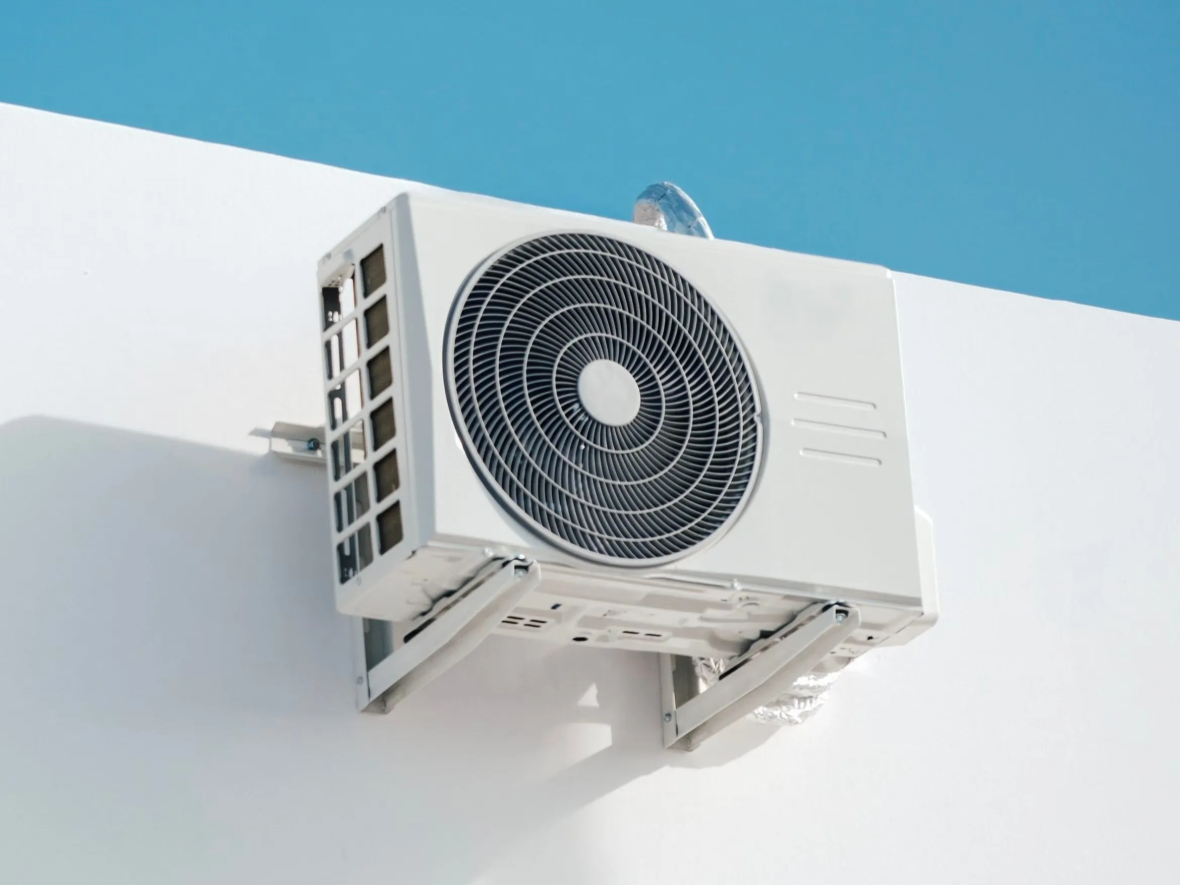 Outdoor mini split unit on metal wall brackets high on a white exterior wall, with its insulated refrigerant line wrapped in foil