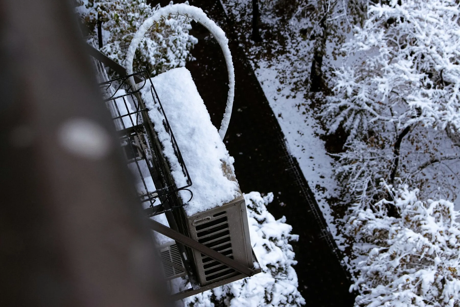 Outdoor mini split unit on an exterior bracket blanketed in snow, with snow-covered trees in the background