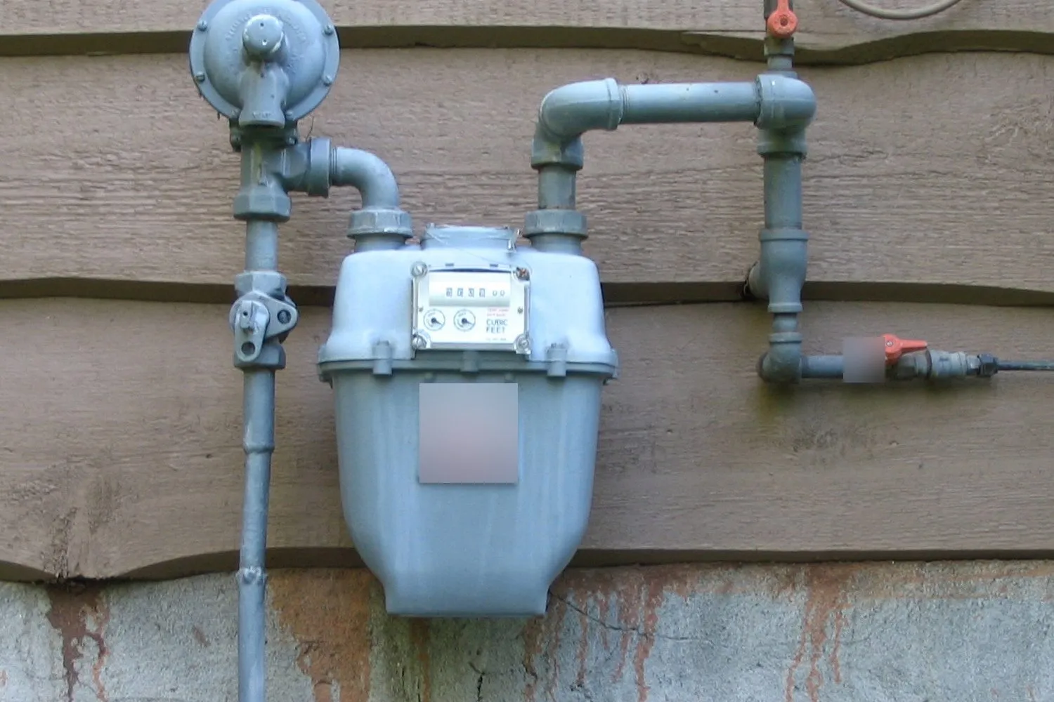 A natural gas meter with its pressure regulator and piping mounted on the wood siding of a house