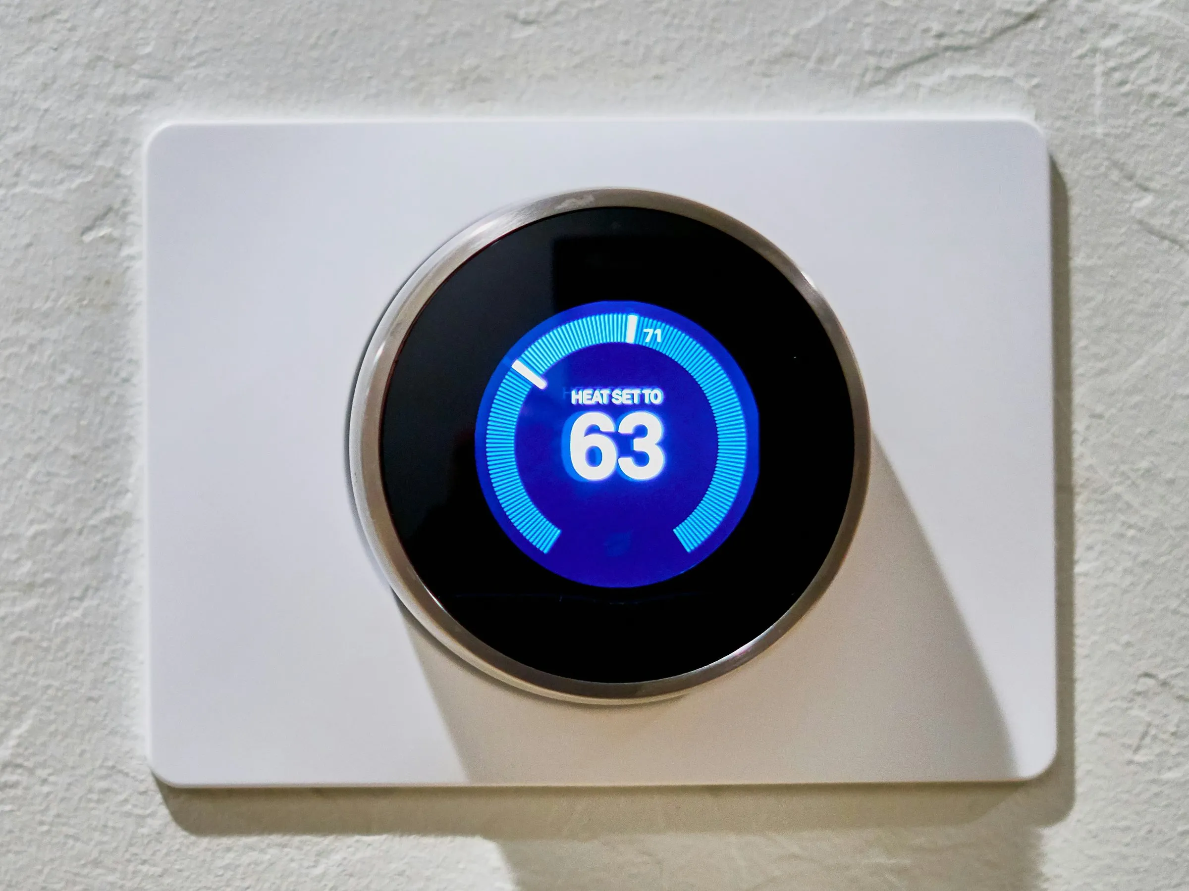 Round digital thermostat on a white wall plate, its screen reading Heat set to 63 with no aux or emergency heat label showing