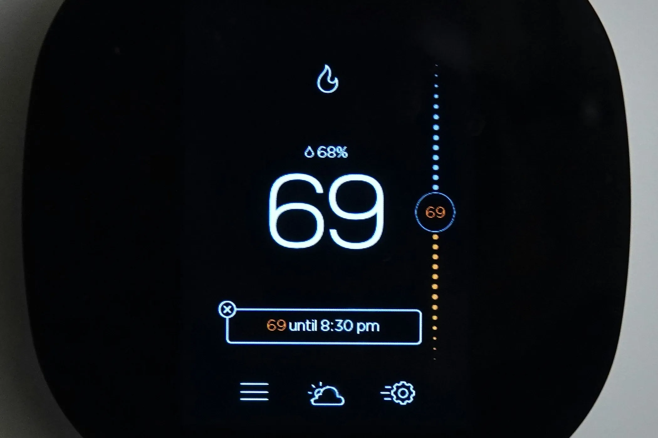 Close-up of a smart thermostat screen in heat mode, with a flame icon, 69 degrees and a hold at 69 until 8:30 pm