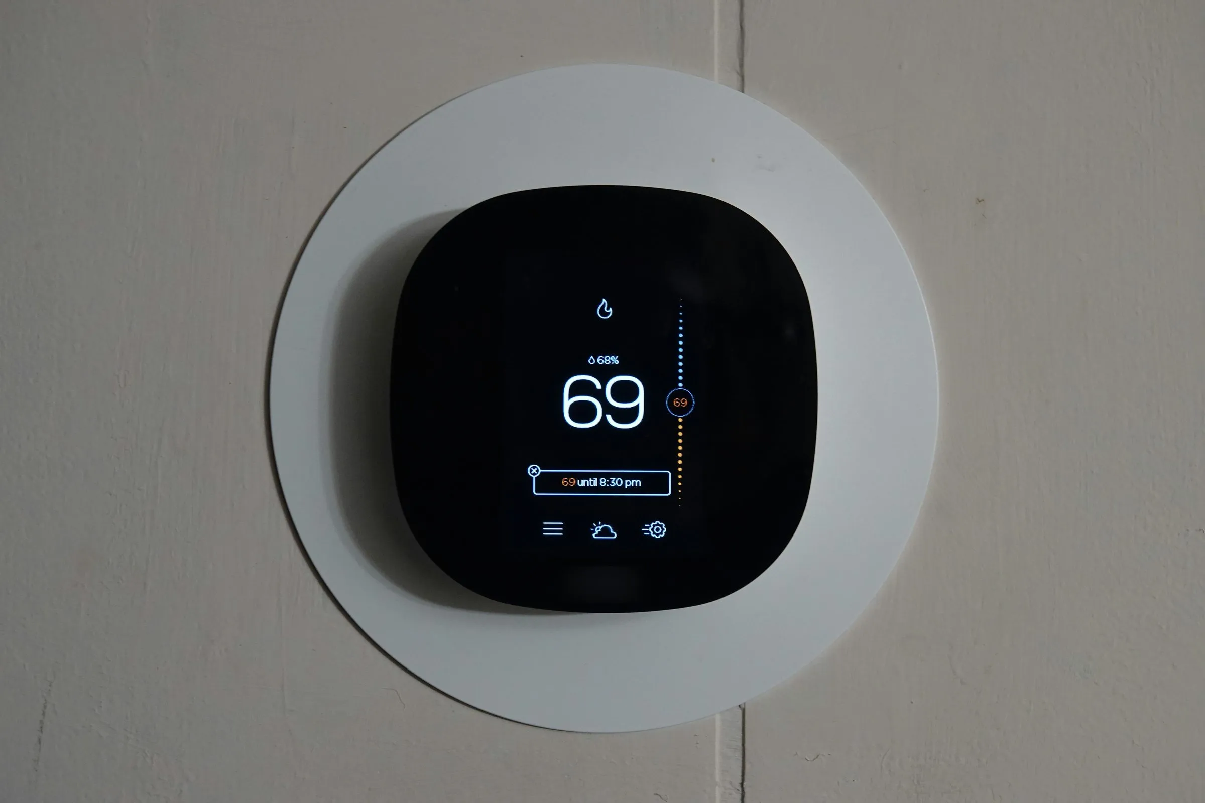 Smart thermostat on a wall in heat mode, its screen showing 69 degrees with a flame icon and 69 until 8:30 pm