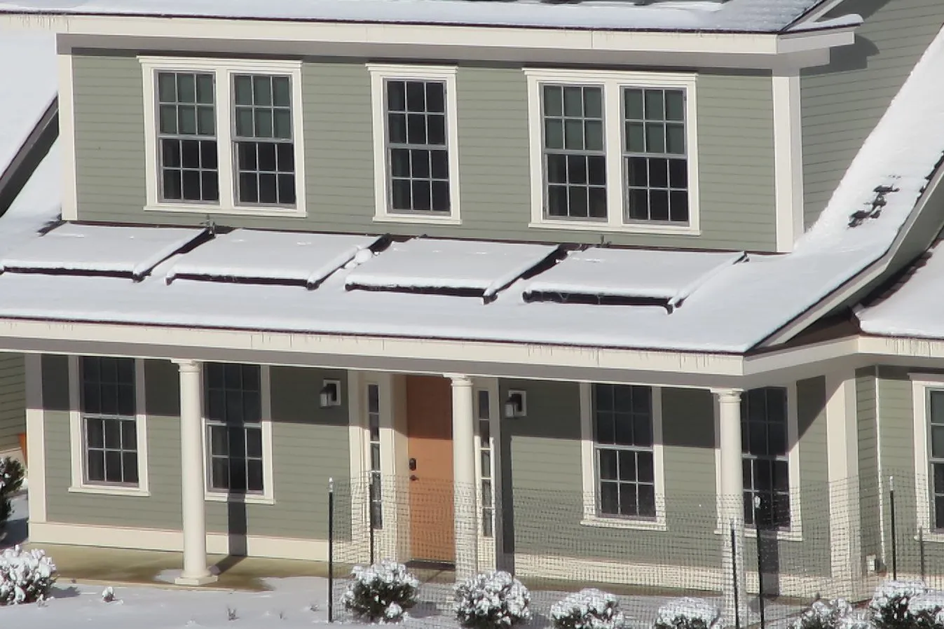 Two-story house with green siding after a snowfall, snow lying on the porch roof and on the shrubs out front on a sunny winter morning