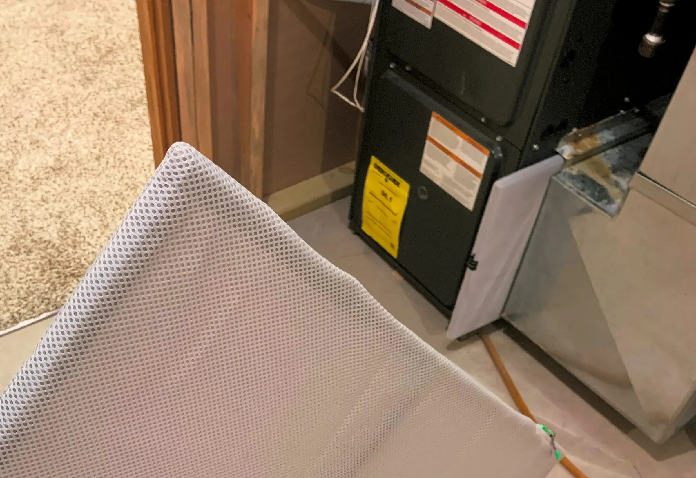 A washable air filter held up beside a gas furnace in a basement, with the filter slot on the side of the return duct