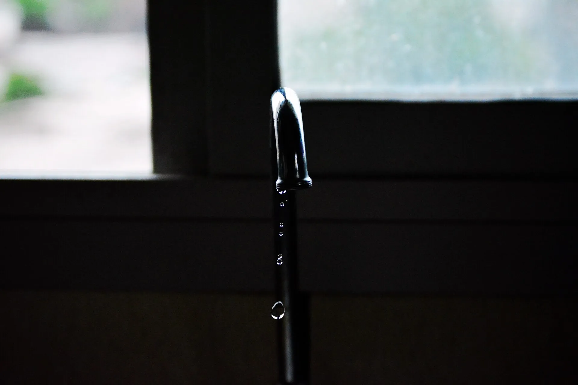 A chrome faucet letting a slow line of water drops fall, with a window behind it